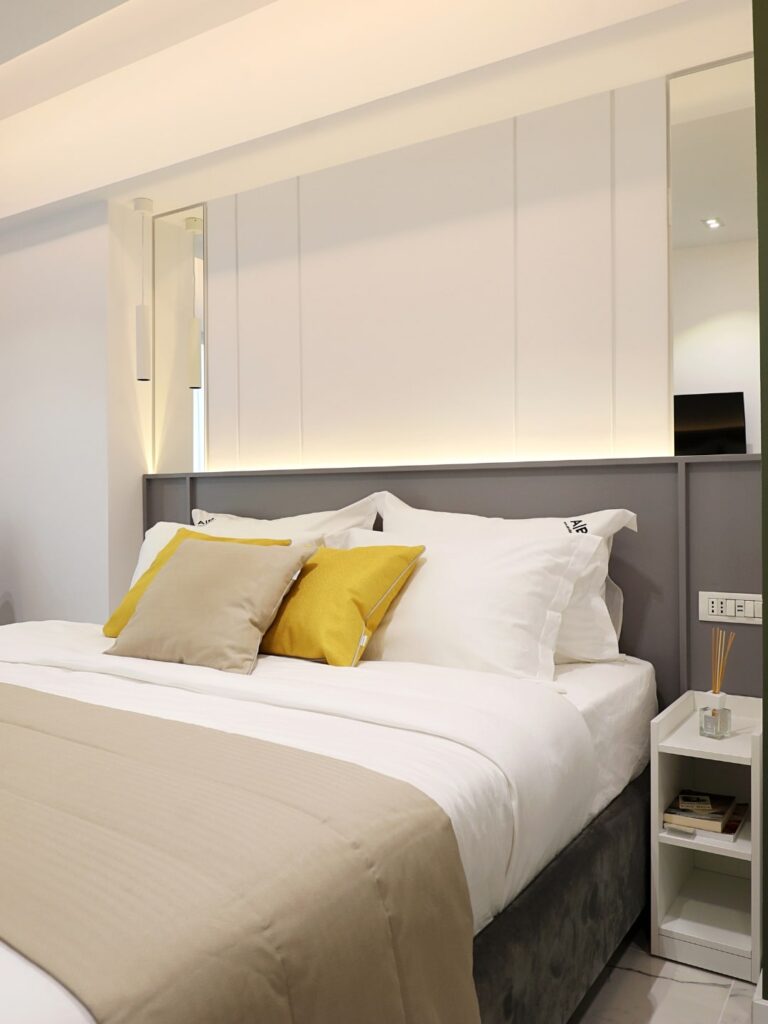 Dormitor minimalist de forma dreptungiulara si un pat mare cu perne galbene si albe.