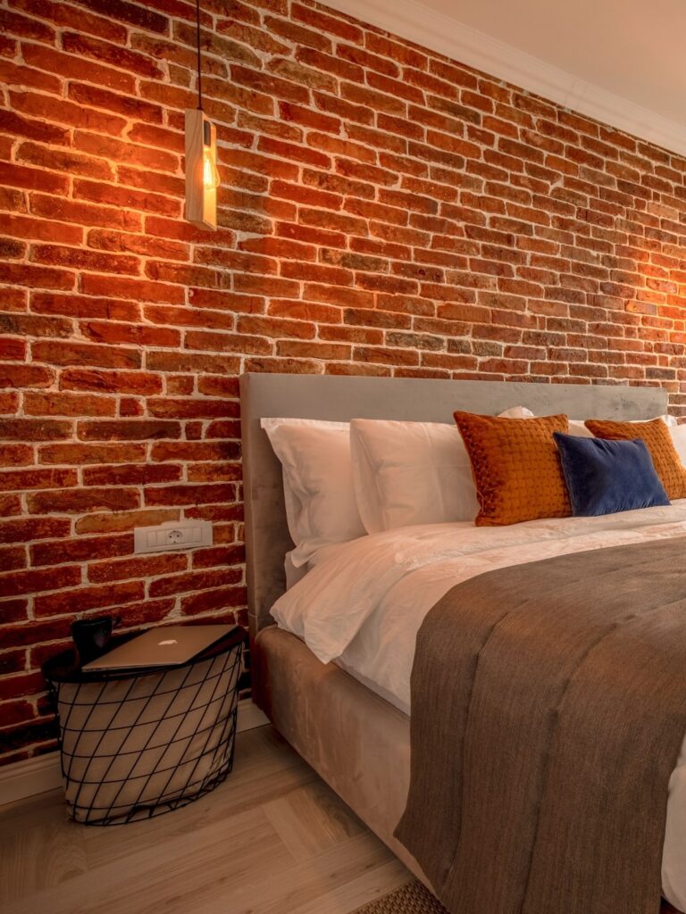 Amenajare dormitor industrial cu un perete din caramida, un pat mare cu multe perne si un bec care ilumineaza camera.