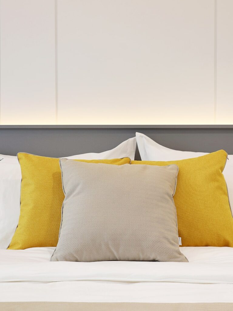 Perne galbene si gri pe un pat de mari dimensiuni in interiorul unui dormitor minimalist.